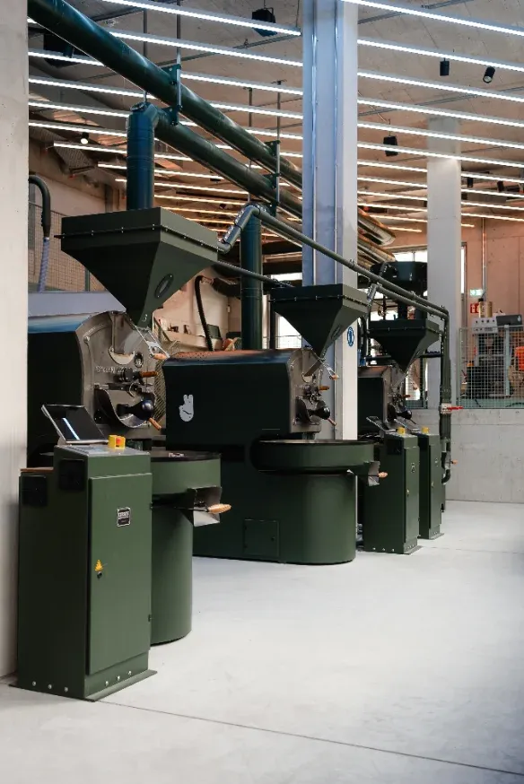 Line of dark-green roasting machines with hoppers and ducting at Alrighty Coffee Roastery in Munich